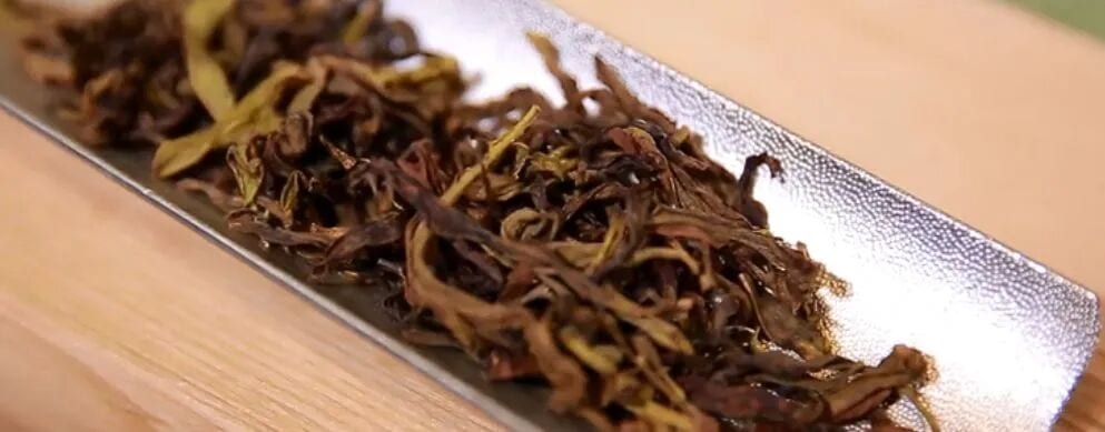 Daun teh kering berwarna gelap dan terpilin di atas nampan teh logam