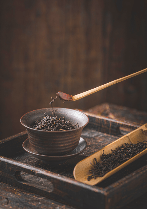 Refined loose tea being placed into a dark ceramic gaiwan with a bamboo scoop